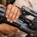 A person wearing MARPAT desert camouflage uniform is handling a disassembled or open grenade launcher, likely an M320 or similar 40mm grenade launcher. The weapon is open/broken down, exposing internal components. Visible markings on the weapon reference '40mm x 46 LV' ammunition caliber and a CAGE code.
