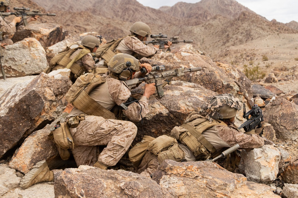 Multiple armed military personnel in desert camouflage uniforms are positioned behind large rocks in a mountainous, arid terrain. They are in tactical firing positions, aiming rifles in a coordinated manner. The setting and equipment suggest a military training exercise or combat operation in a desert environment.