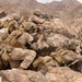 Multiple armed military personnel in desert camouflage uniforms are positioned behind large rocks in a mountainous, arid terrain. They are in tactical firing positions, aiming rifles in a coordinated manner. The setting and equipment suggest a military training exercise or combat operation in a desert environment.