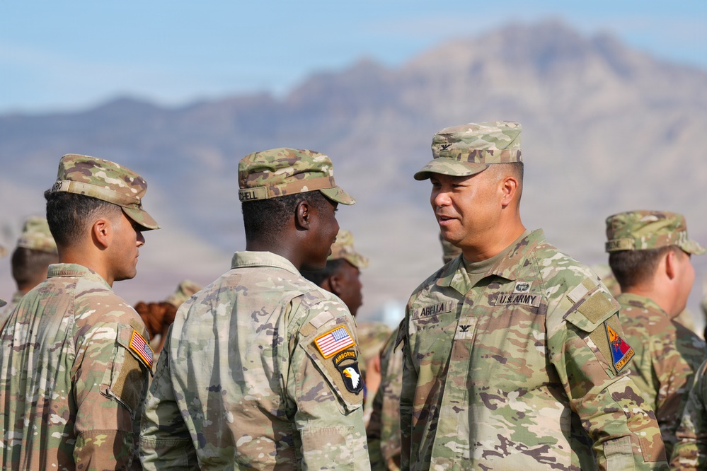 A group of U.S. Army soldiers in OCP (Operational Camouflage Pattern) uniforms are gathered outdoors in a desert environment with mountains visible in the background. A senior officer, identified by his name tape 'ABELLA' and colonel rank insignia (eagle), appears to be speaking with junior soldiers. At least one soldier displays a 101st Airborne Division patch on their left shoulder.