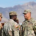 A group of U.S. Army soldiers in OCP (Operational Camouflage Pattern) uniforms are gathered outdoors in a desert environment with mountains visible in the background. A senior officer, identified by his name tape 'ABELLA' and colonel rank insignia (eagle), appears to be speaking with junior soldiers. At least one soldier displays a 101st Airborne Division patch on their left shoulder.