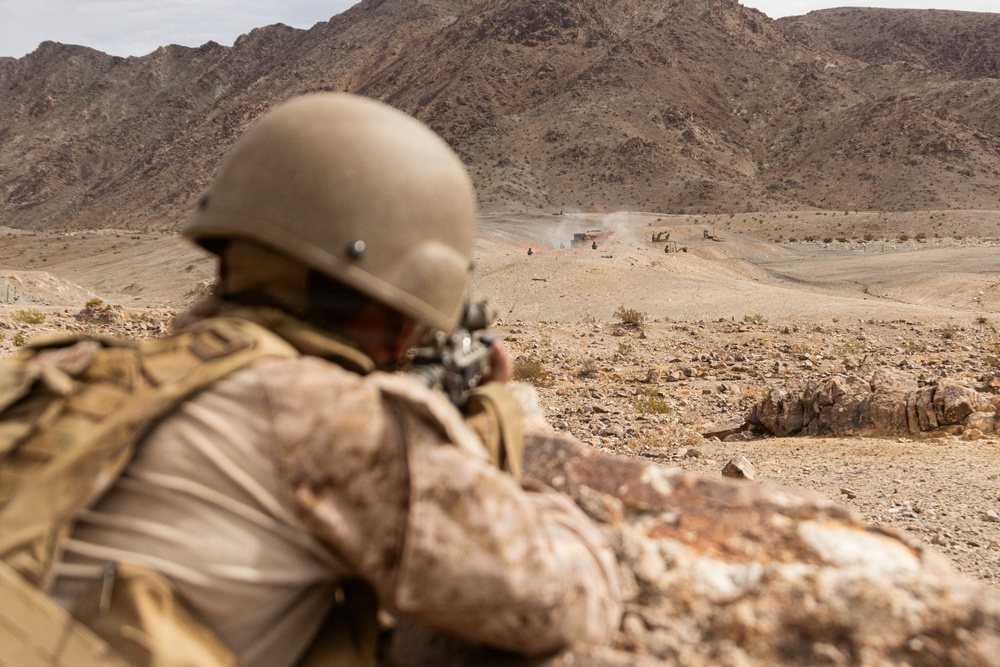 A soldier in desert camouflage uniform and helmet is in a prone firing position, aiming a rifle toward a distant target area in an arid, rocky desert landscape. Smoke or dust is visible in the mid-ground near what appears to be structures or vehicles, suggesting an active training exercise or engagement. The mountainous terrain and sandy environment are consistent with a desert training range.