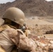 A soldier in desert camouflage uniform and helmet is in a prone firing position, aiming a rifle toward a distant target area in an arid, rocky desert landscape. Smoke or dust is visible in the mid-ground near what appears to be structures or vehicles, suggesting an active training exercise or engagement. The mountainous terrain and sandy environment are consistent with a desert training range.