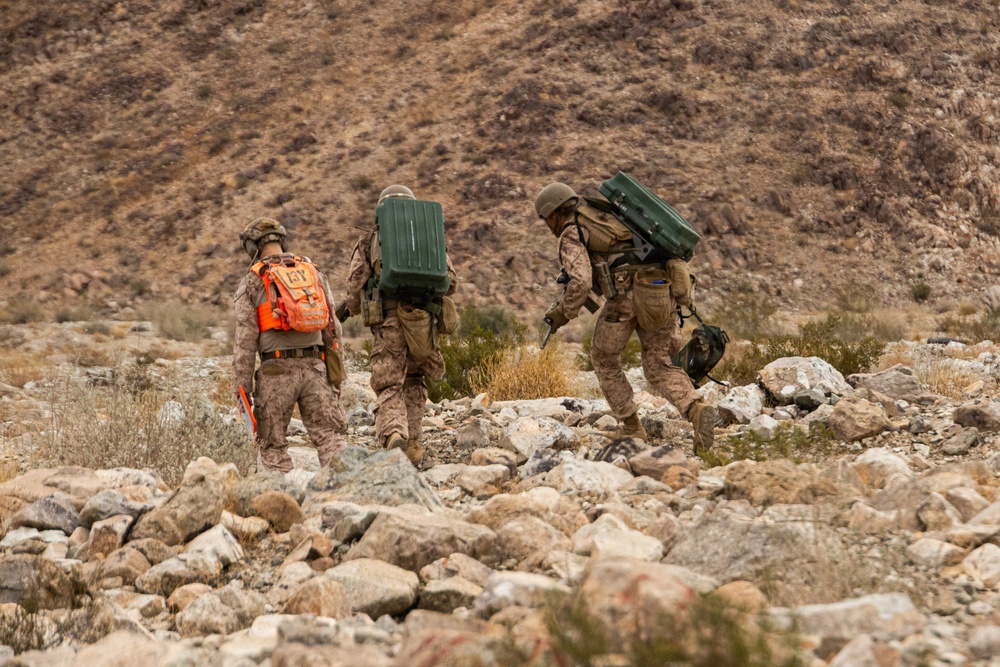 Three military personnel in desert camouflage uniforms are traversing rocky terrain in an arid, mountainous environment. Two of the soldiers are carrying large green military equipment cases on their backs, while one wears a distinctive bright orange pack. The setting appears to be a desert training exercise or field operation.