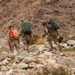 Three military personnel in desert camouflage uniforms are traversing rocky terrain in an arid, mountainous environment. Two of the soldiers are carrying large green military equipment cases on their backs, while one wears a distinctive bright orange pack. The setting appears to be a desert training exercise or field operation.