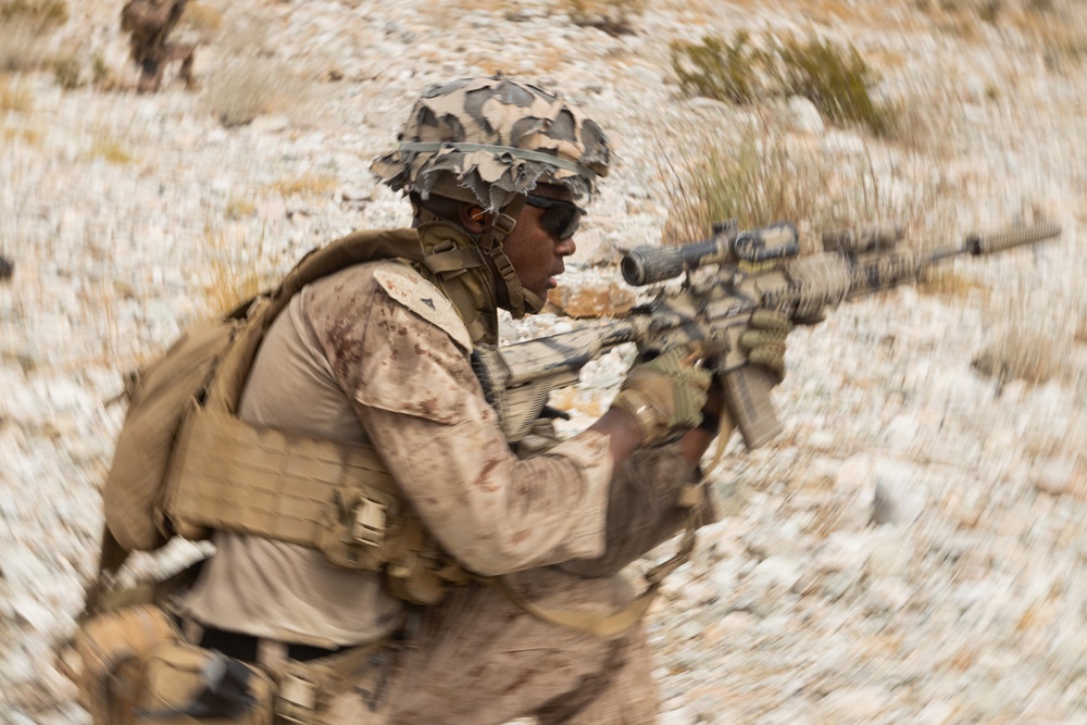 A soldier in desert camouflage uniform is captured in motion while aiming a rifle equipped with an optical scope. The image uses a radial blur/zoom effect centered on the subject, creating a sense of dynamic movement. A second figure is faintly visible in the background on elevated terrain.