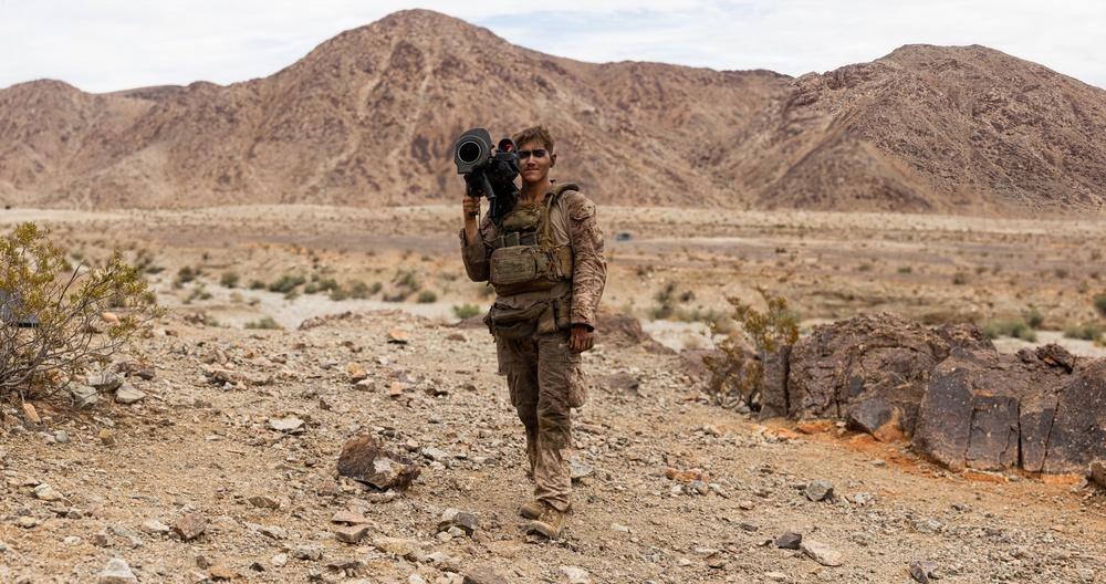 A soldier in desert camouflage uniform carries a shoulder-mounted weapon system while walking through an arid desert terrain. The individual is equipped with tactical gear including a chest rig and appears to be on a training exercise or patrol. Rocky mountains and sparse desert vegetation form the background.