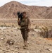 A soldier in desert camouflage uniform carries a shoulder-mounted weapon system while walking through an arid desert terrain. The individual is equipped with tactical gear including a chest rig and appears to be on a training exercise or patrol. Rocky mountains and sparse desert vegetation form the background.
