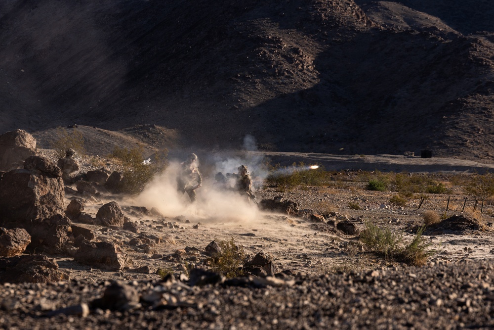 The image shows military personnel in desert terrain firing what appears to be a shoulder-launched rocket or missile system. A visible projectile trail and dust cloud are captured mid-launch in a rocky, arid landscape. The scene appears to be either a live-fire training exercise or combat operation based on the controlled positioning of the personnel.