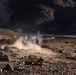 The image shows military personnel in desert terrain firing what appears to be a shoulder-launched rocket or missile system. A visible projectile trail and dust cloud are captured mid-launch in a rocky, arid landscape. The scene appears to be either a live-fire training exercise or combat operation based on the controlled positioning of the personnel.