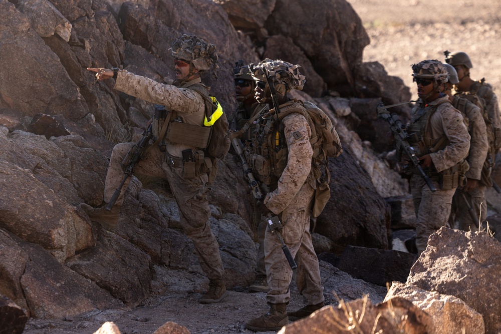 A group of military personnel in desert camouflage uniforms are positioned among large rocks in an arid, rocky terrain. One soldier in the foreground is pointing in a direction, appearing to give tactical guidance to the others. The scene appears to be a military training exercise or field operation, indicated by the yellow training markers visible on some personnel.