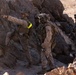 A group of military personnel in desert camouflage uniforms are positioned among large rocks in an arid, rocky terrain. One soldier in the foreground is pointing in a direction, appearing to give tactical guidance to the others. The scene appears to be a military training exercise or field operation, indicated by the yellow training markers visible on some personnel.