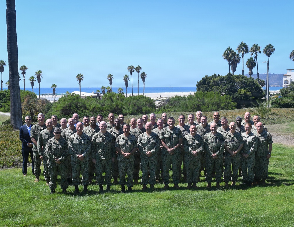 A large group of approximately 35-40 military personnel, predominantly wearing U.S. Navy NWU Type III (woodland camouflage) uniforms, pose for a formal group photograph outdoors. They are arranged in two to three rows on a grass lawn with palm trees, ocean, and a sandy beach visible in the background, suggesting a coastal military installation likely in Southern California. One individual on the far left is wearing civilian business attire.