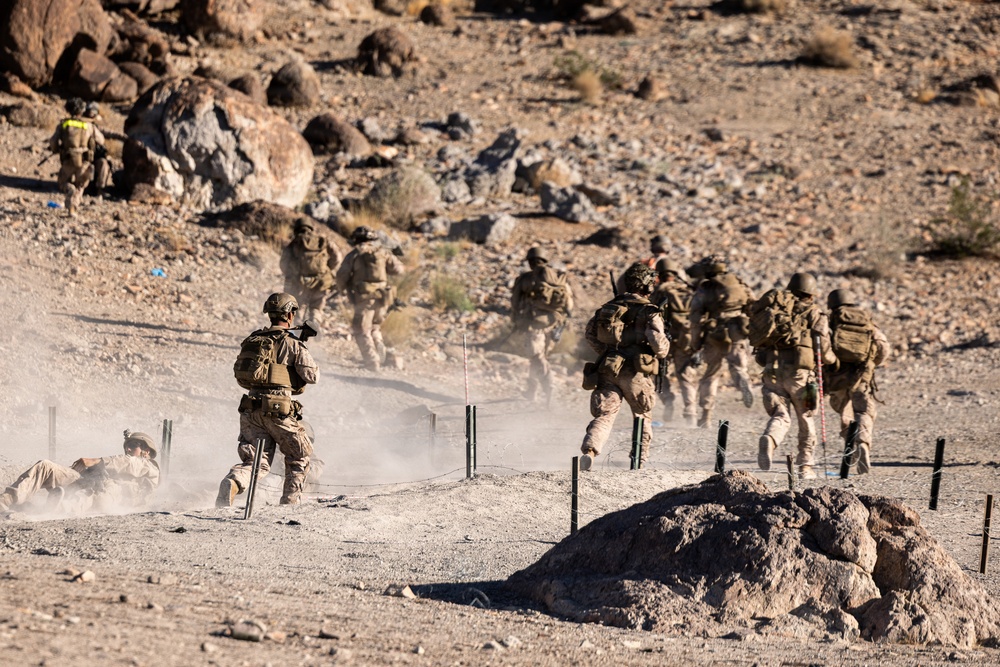 A group of military personnel in desert camouflage uniforms are conducting what appears to be a tactical training exercise in an arid, rocky desert environment. Multiple soldiers are advancing forward while at least one individual is prone on the ground, suggesting a live-fire or assault drill. The terrain features sandy soil, scattered rocks, and boundary markers including posts with wire or rope.