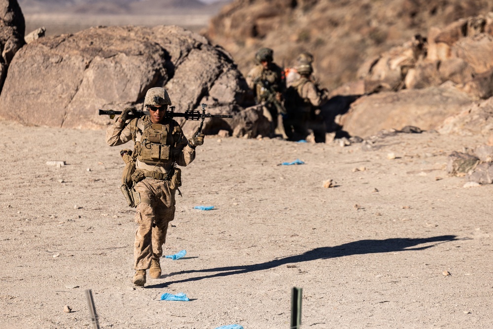 A military service member in desert camouflage uniform is walking forward while carrying a large weapon system across their shoulders in an arid, rocky terrain. In the background, two additional personnel are positioned near large boulders, appearing to be in a tactical formation. The setting and equipment suggest a military training exercise in a desert environment.