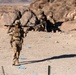 A military service member in desert camouflage uniform is walking forward while carrying a large weapon system across their shoulders in an arid, rocky terrain. In the background, two additional personnel are positioned near large boulders, appearing to be in a tactical formation. The setting and equipment suggest a military training exercise in a desert environment.