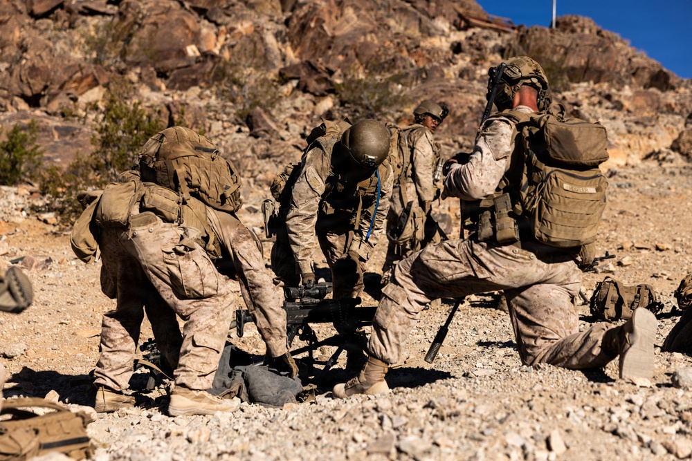 Multiple military personnel in desert camouflage uniforms are working together to set up or operate a crew-served weapon system in an arid, rocky terrain. The soldiers are wearing full combat gear including tactical vests, helmets, and backpacks. The setting appears to be a training exercise in a desert environment, likely in the American Southwest based on the terrain and rock formations.