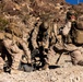 Multiple military personnel in desert camouflage uniforms are working together to set up or operate a crew-served weapon system in an arid, rocky terrain. The soldiers are wearing full combat gear including tactical vests, helmets, and backpacks. The setting appears to be a training exercise in a desert environment, likely in the American Southwest based on the terrain and rock formations.