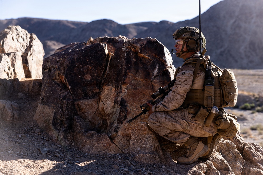 A uniformed military service member in desert camouflage is positioned behind a large boulder in an arid, rocky desert environment. The soldier is equipped with full combat gear including a helmet with communications equipment, body armor, a backpack, and is holding a rifle. The setting appears to be a training exercise or operational patrol in a desert terrain with mountains visible in the background.