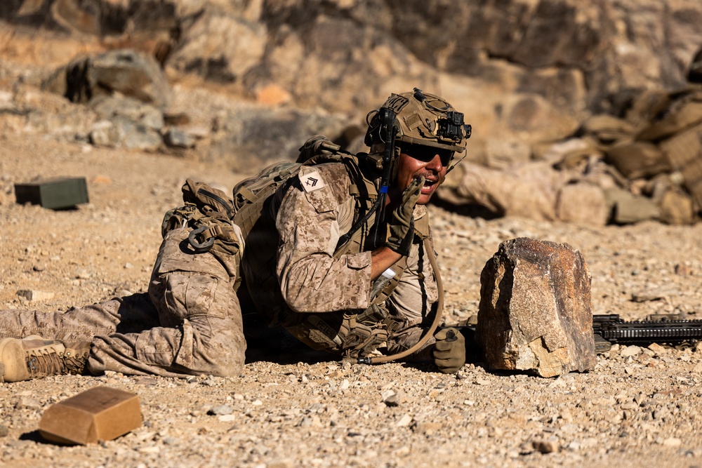 A soldier in desert camouflage uniform is in a prone position on arid, rocky terrain, appearing to shout or communicate while taking cover behind a large rock. The soldier is fully equipped with tactical gear including a helmet with night-vision mount, backpack, gloves, and a rifle visible to the right. The setting appears to be a desert environment, consistent with a training exercise or combat operation.