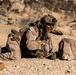 A soldier in desert camouflage uniform is in a prone position on arid, rocky terrain, appearing to shout or communicate while taking cover behind a large rock. The soldier is fully equipped with tactical gear including a helmet with night-vision mount, backpack, gloves, and a rifle visible to the right. The setting appears to be a desert environment, consistent with a training exercise or combat operation.