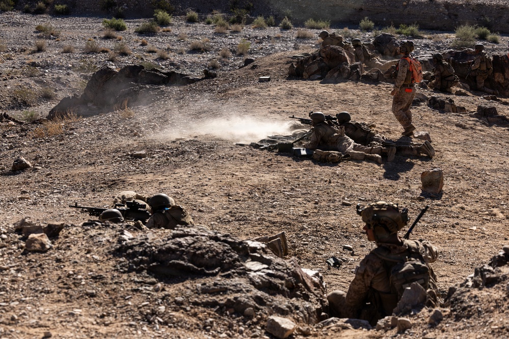The image depicts a military training exercise in an arid, rocky desert environment. Multiple soldiers in desert camouflage uniforms are positioned in various tactical stances including prone firing positions and covered positions behind rocks. A distinctive orange-vested individual, likely a range safety officer or observer, is standing upright among the group.