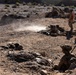 The image depicts a military training exercise in an arid, rocky desert environment. Multiple soldiers in desert camouflage uniforms are positioned in various tactical stances including prone firing positions and covered positions behind rocks. A distinctive orange-vested individual, likely a range safety officer or observer, is standing upright among the group.