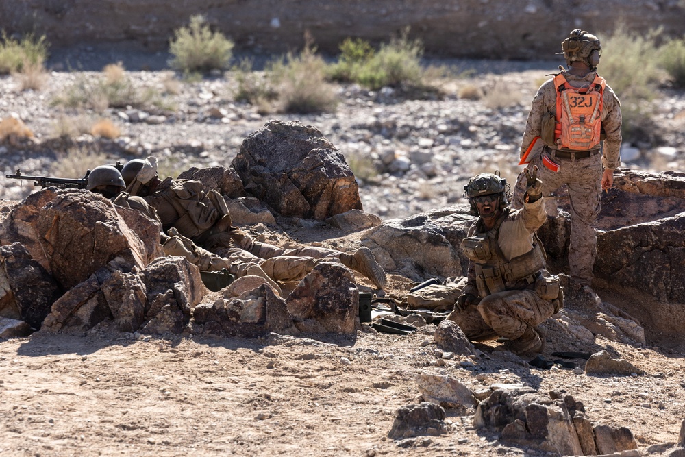 The image shows military personnel in desert camouflage uniforms conducting what appears to be a training exercise in an arid, rocky terrain. One individual wearing a bright orange safety vest marked '32J' stands upright, likely serving as a safety observer or evaluator. Two other personnel are positioned low among the rocks, one appearing to be in a firing position with a weapon.