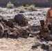 The image shows military personnel in desert camouflage uniforms conducting what appears to be a training exercise in an arid, rocky terrain. One individual wearing a bright orange safety vest marked '32J' stands upright, likely serving as a safety observer or evaluator. Two other personnel are positioned low among the rocks, one appearing to be in a firing position with a weapon.