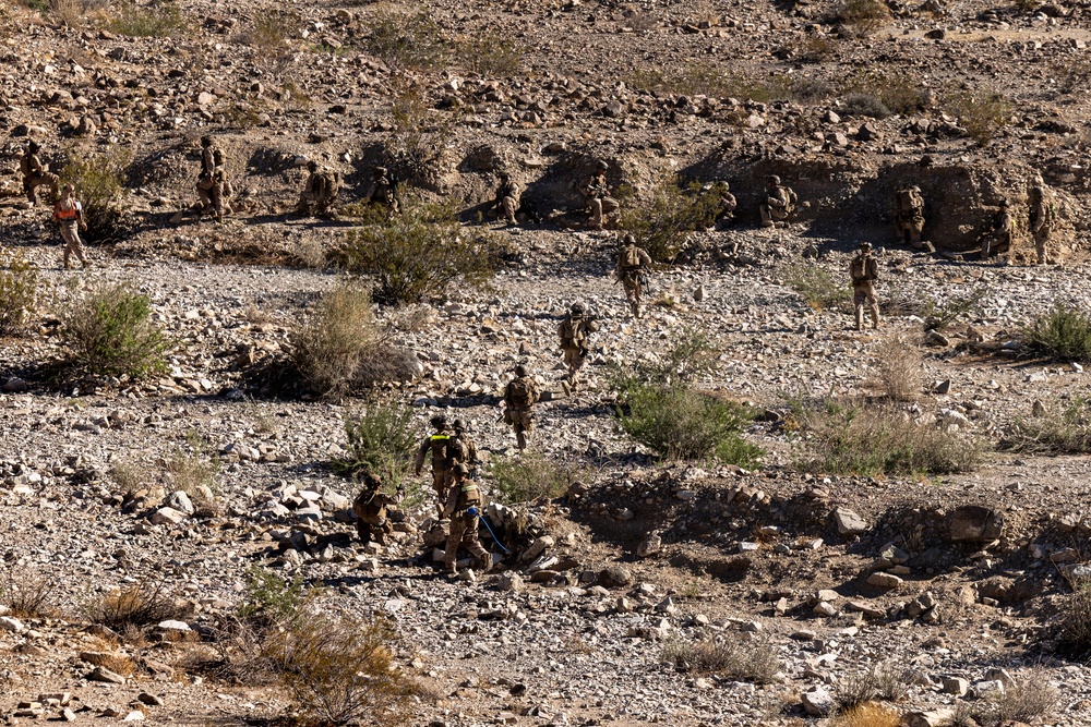 The image shows a group of military personnel conducting a patrol or training exercise across a rocky, arid desert terrain. The soldiers are spread out in a dispersed formation moving uphill through a dry, scrub-covered landscape. The environment appears consistent with a desert training area such as those found in the American Southwest.