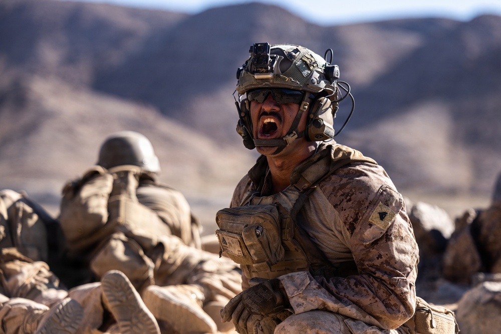 A soldier in full combat gear is shouting with mouth wide open in an arid, desert environment. The individual is equipped with advanced tactical equipment including a helmet-mounted camera, night vision mount, and communications headset. Additional personnel in similar desert camouflage uniforms are visible in the blurred background.
