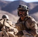 A soldier in full combat gear is shouting with mouth wide open in an arid, desert environment. The individual is equipped with advanced tactical equipment including a helmet-mounted camera, night vision mount, and communications headset. Additional personnel in similar desert camouflage uniforms are visible in the blurred background.