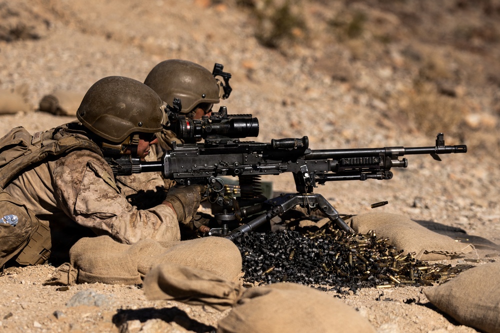 Two military personnel in desert camouflage uniforms and tactical helmets are operating a crew-served machine gun from a prone position behind sandbag fortifications. The weapon appears to be an M240 machine gun mounted on a bipod, with a large accumulation of spent brass and linked ammunition casings visible in the foreground. The setting appears to be an arid, desert training range environment.