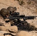 Two military personnel in desert camouflage uniforms and tactical helmets are operating a crew-served machine gun from a prone position behind sandbag fortifications. The weapon appears to be an M240 machine gun mounted on a bipod, with a large accumulation of spent brass and linked ammunition casings visible in the foreground. The setting appears to be an arid, desert training range environment.