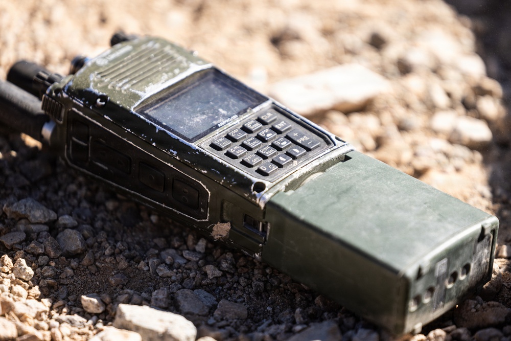 A heavily worn military-grade handheld radio transceiver lies on rocky, gravelly ground. The device is olive drab/green in color, showing significant wear, scratches, and dirt consistent with field use. It features a keypad, small display screen, antenna, and a large battery pack attached to the rear.