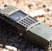 A heavily worn military-grade handheld radio transceiver lies on rocky, gravelly ground. The device is olive drab/green in color, showing significant wear, scratches, and dirt consistent with field use. It features a keypad, small display screen, antenna, and a large battery pack attached to the rear.