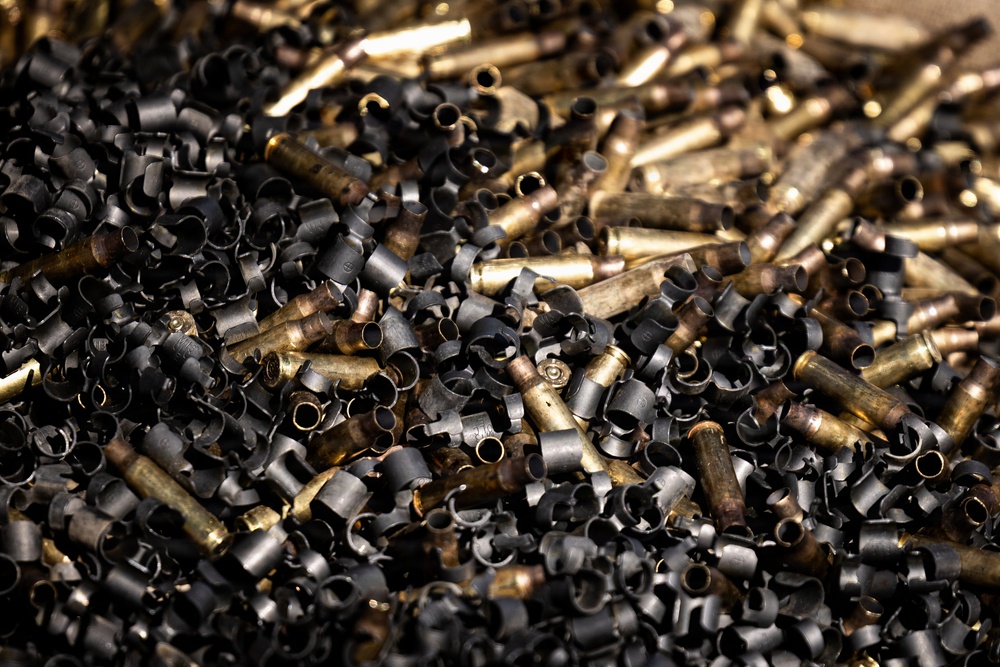 The image shows a large pile of spent brass bullet casings mixed with dark metal shavings or curled metal debris. The casings appear to be of varying calibers, predominantly rifle-sized, in various states of oxidation ranging from bright brass to darkened patina. This appears to be a collection of recycled or discarded ammunition components, likely from a shooting range or metal recycling facility.