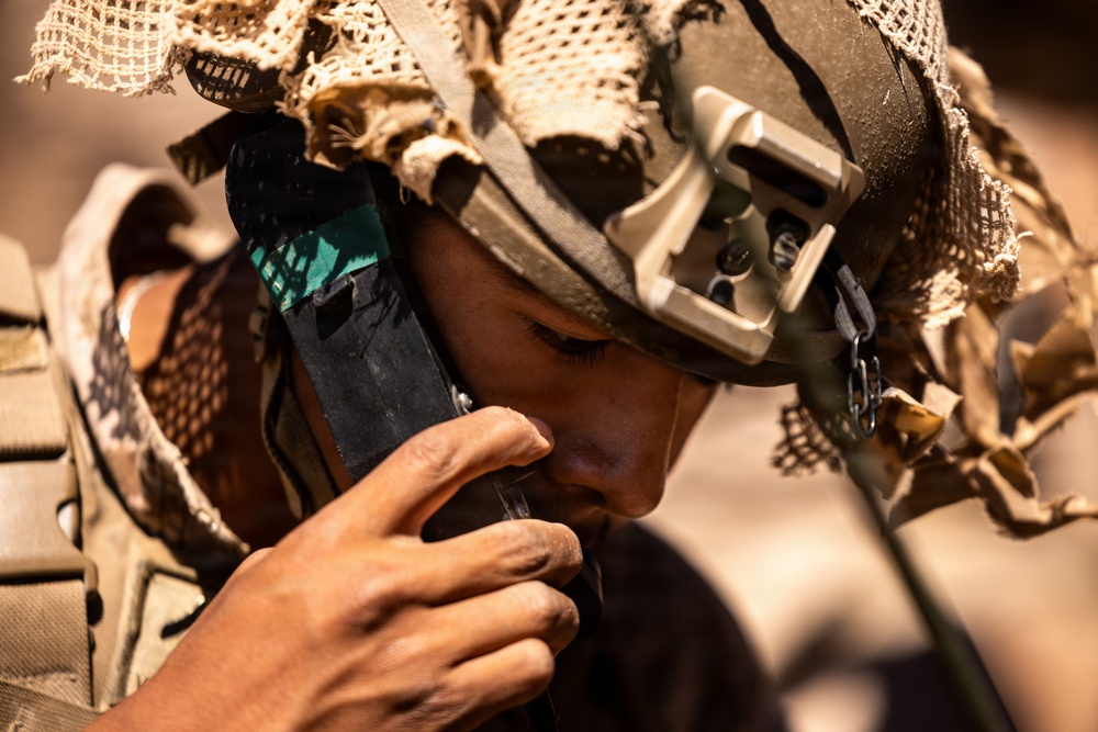 A soldier in full tactical gear is drinking water from a hydration tube or water pouch in what appears to be a desert or arid environment. The individual is wearing a camouflage helmet with netting (ghillie-style cover) and a night-vision goggle mount bracket. The close-up shot captures fine details of the equipment and the act of hydration during what appears to be field operations.