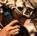 A soldier in full tactical gear is drinking water from a hydration tube or water pouch in what appears to be a desert or arid environment. The individual is wearing a camouflage helmet with netting (ghillie-style cover) and a night-vision goggle mount bracket. The close-up shot captures fine details of the equipment and the act of hydration during what appears to be field operations.