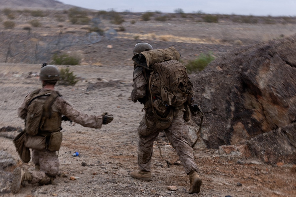 Two military personnel in desert camouflage uniforms are moving through an arid, rocky terrain. Both are wearing helmets and carrying tactical gear and backpacks, suggesting a field training exercise or operational movement. The environment appears to be a desert or semi-arid region with sparse vegetation and rocky outcroppings.