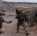 Two military personnel in desert camouflage uniforms are moving through an arid, rocky terrain. Both are wearing helmets and carrying tactical gear and backpacks, suggesting a field training exercise or operational movement. The environment appears to be a desert or semi-arid region with sparse vegetation and rocky outcroppings.