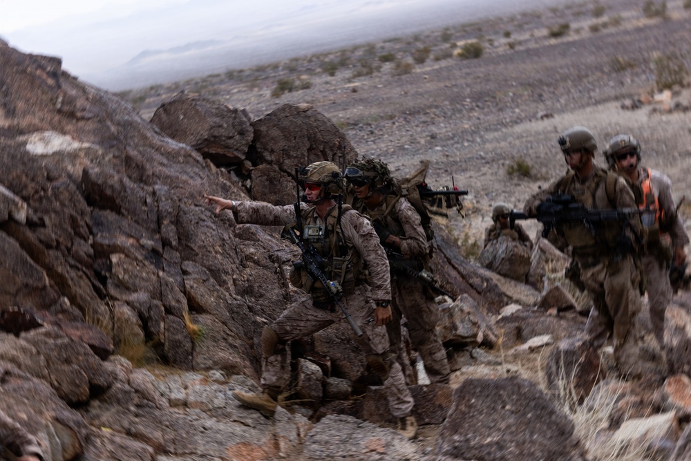 A group of military personnel in full combat gear are moving through a rocky desert terrain, navigating a narrow rocky passage. One soldier in the foreground is pointing in a direction, appearing to give guidance or signal to the others. The arid, mountainous landscape and camouflage uniforms suggest a training exercise or field operation.