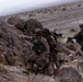 A group of military personnel in full combat gear are moving through a rocky desert terrain, navigating a narrow rocky passage. One soldier in the foreground is pointing in a direction, appearing to give guidance or signal to the others. The arid, mountainous landscape and camouflage uniforms suggest a training exercise or field operation.