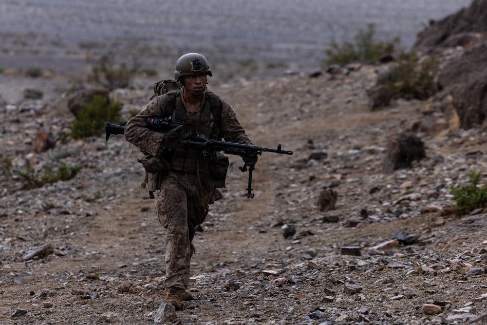 A soldier in desert camouflage uniform and combat helmet is running across rocky, arid terrain while carrying a light machine gun with a bipod deployed. The environment appears to be a desert or semi-arid training area, with sparse vegetation and rocky ground. The overcast, hazy conditions suggest either rain or dust in the atmosphere.