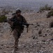 A soldier in desert camouflage uniform and combat helmet is running across rocky, arid terrain while carrying a light machine gun with a bipod deployed. The environment appears to be a desert or semi-arid training area, with sparse vegetation and rocky ground. The overcast, hazy conditions suggest either rain or dust in the atmosphere.