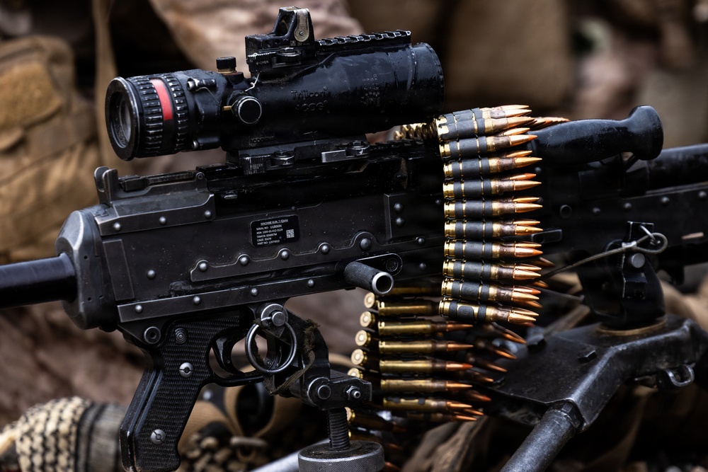A close-up photograph of a belt-fed machine gun, identifiable as an M249 SAW or similar light machine gun variant, equipped with a Trijicon ACOG optical sight and a miniature red dot sight mounted on top. A linked ammunition belt with brass-cased, copper-jacketed rounds is loaded into the weapon's feed tray. The weapon appears to be in a field or operational environment, resting on military equipment.