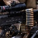 A close-up photograph of a belt-fed machine gun, identifiable as an M249 SAW or similar light machine gun variant, equipped with a Trijicon ACOG optical sight and a miniature red dot sight mounted on top. A linked ammunition belt with brass-cased, copper-jacketed rounds is loaded into the weapon's feed tray. The weapon appears to be in a field or operational environment, resting on military equipment.