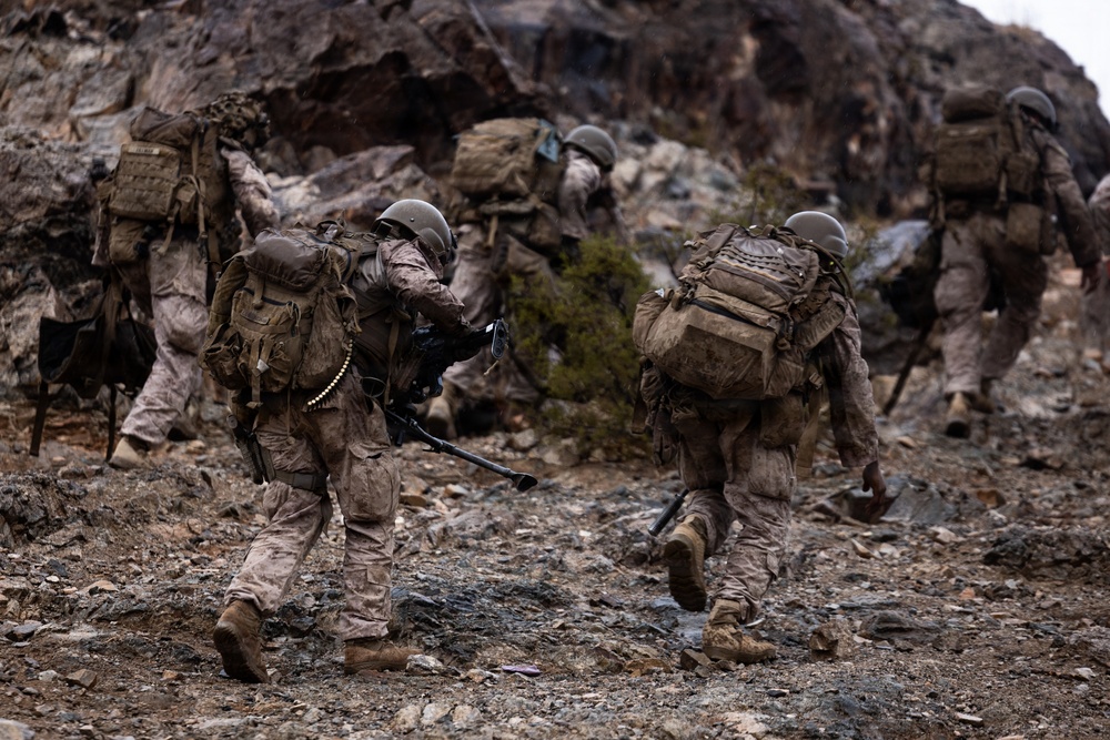 A group of heavily equipped military personnel in desert camouflage uniforms are moving through a rocky, mountainous terrain. The soldiers are carrying large backpacks, weapons, and ammunition belts, suggesting a patrol or tactical movement exercise. The environment appears arid and rugged, consistent with a desert or semi-arid mountain region.