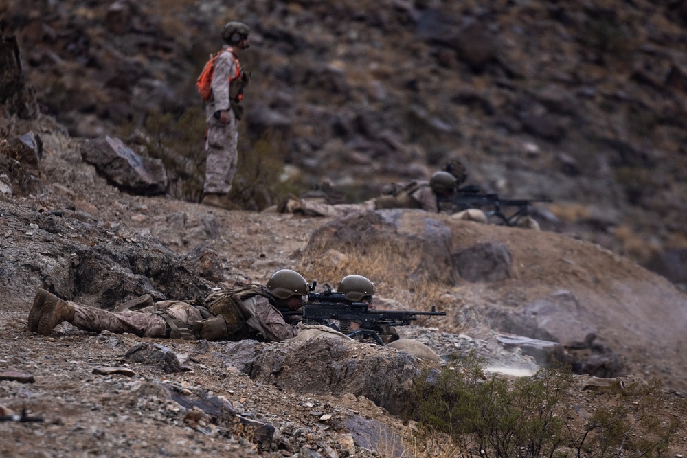 The image depicts military personnel conducting what appears to be a live-fire training exercise on a rocky, arid hillside terrain. Two soldiers in the foreground are in a prone firing position operating a light machine gun, while a third soldier stands upright in the background wearing a high-visibility orange vest, likely serving as a safety observer or range control officer. A fourth personnel is visible in the mid-ground also in a firing position.