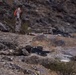 The image depicts military personnel conducting what appears to be a live-fire training exercise on a rocky, arid hillside terrain. Two soldiers in the foreground are in a prone firing position operating a light machine gun, while a third soldier stands upright in the background wearing a high-visibility orange vest, likely serving as a safety observer or range control officer. A fourth personnel is visible in the mid-ground also in a firing position.